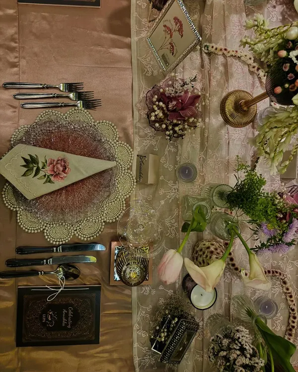 A detailed flat lay of a table setting for a very special birthday dinner. The combination of a lace overlay, floral-printed napkin, and personalized menu book creates a romantic and sophisticated atmosphere.