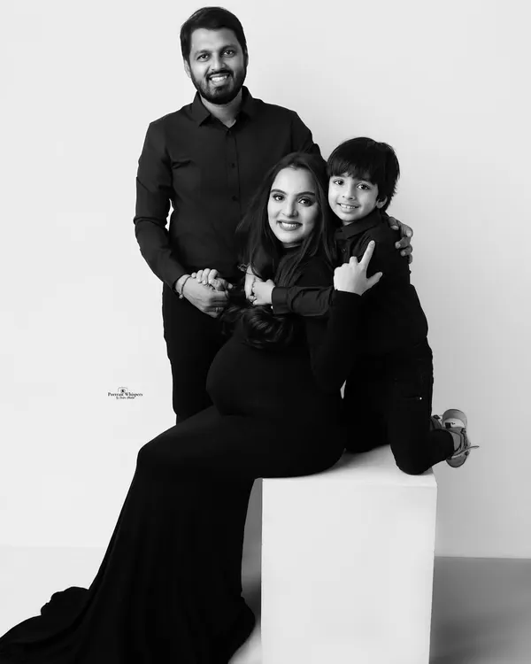 A joyful black and white family portrait. The mother-to-be is seated while her husband and son embrace her, all sharing a beautiful, happy moment.