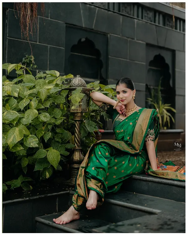A stunning portrait of a Maharashtrian bride in a rich green Paithani saree. Her graceful pose on the steps, surrounded by greenery, highlights her timeless elegance.