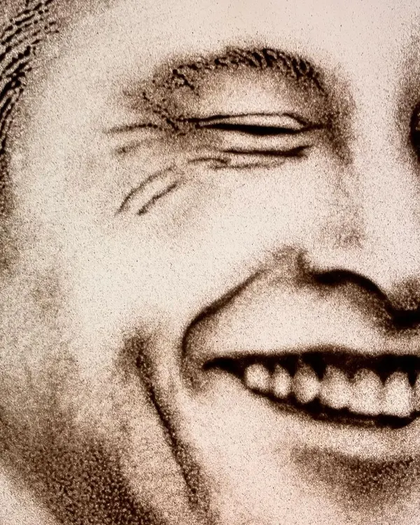 Can you guess who this is? A close-up detail of a sand portrait capturing the iconic smile of a visionary. The texture of the sand adds a unique depth to the expression.