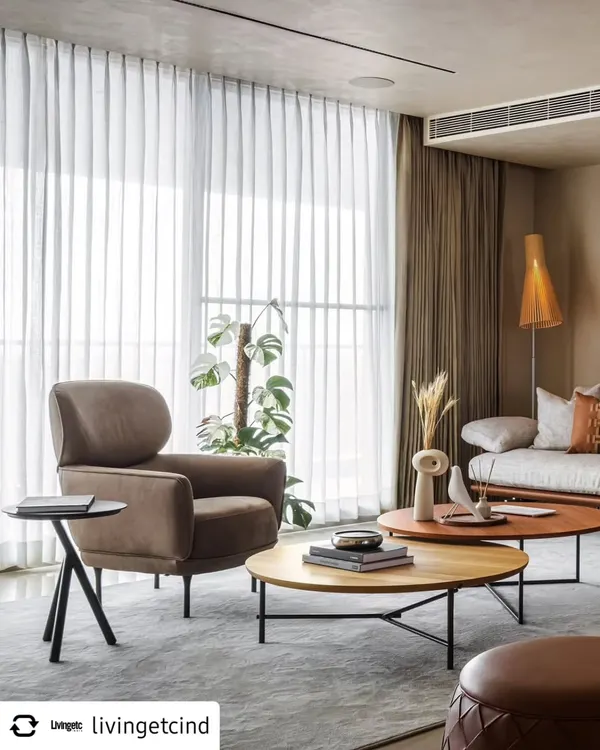 In the living area of Celadon, we layered textures and neutral tones to create a calm, inviting atmosphere. The soft brown armchair and nested wooden coffee tables sit before sheer curtains that diffuse natural light, enhancing the connection to the lush greenery just outside.