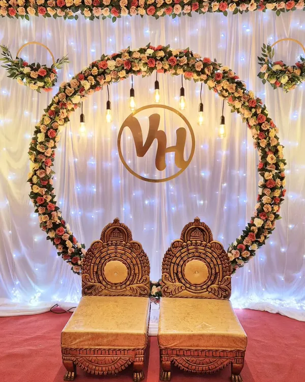 A stunning engagement stage featuring a large ring made of artificial roses, illuminated by hanging Edison bulbs. A custom gold monogram and traditional seating complete this classic and elegant look.