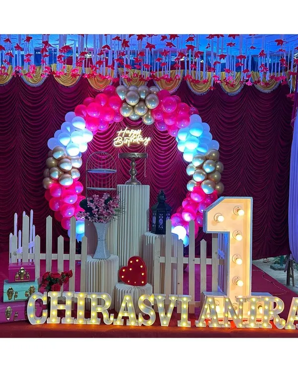 A festive first birthday stage with a circular balloon arch in red, white, and gold. The setup is personalized with the child's name in large marquee letters.