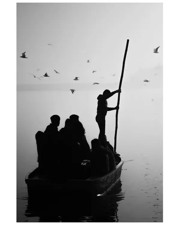 A black and white silhouette of a boatman steering his vessel through the foggy waters of the Yamuna. The image has a timeless, serene quality, capturing the quiet dedication of his work.