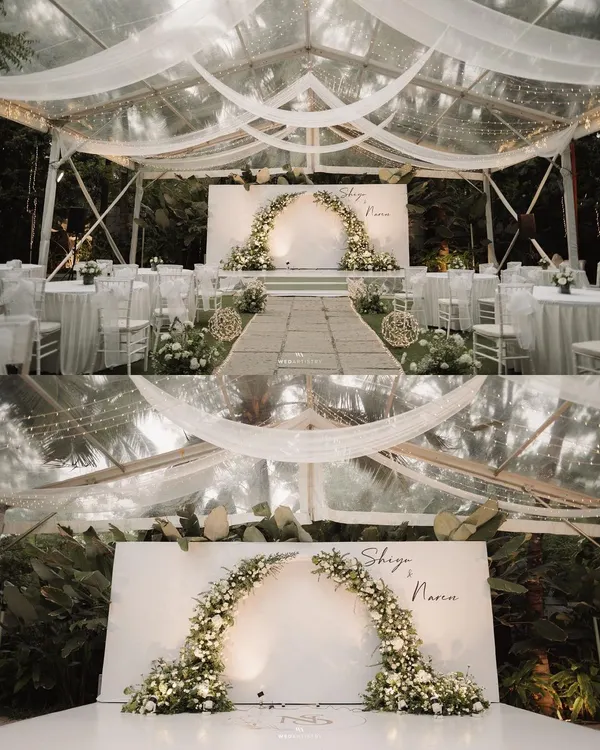 A serene and intimate wedding reception setup inside a transparent tent. The decor is all-white, featuring a beautiful floral arch, clean tablescapes, and elegant draping that creates a dreamy, romantic atmosphere.