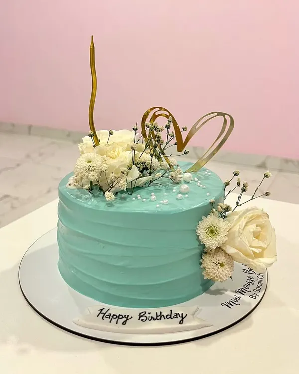 A slice of floral heaven. This elegant birthday cake has a textured teal buttercream finish and is decorated with fresh white flowers, baby's breath, and a delicate gold heart topper.