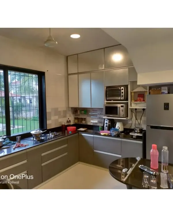 This L-shaped kitchen features laminate cabinets with sleek profile handles and a dark granite countertop. The layout is designed for practicality, with the refrigerator and microwave easily accessible.