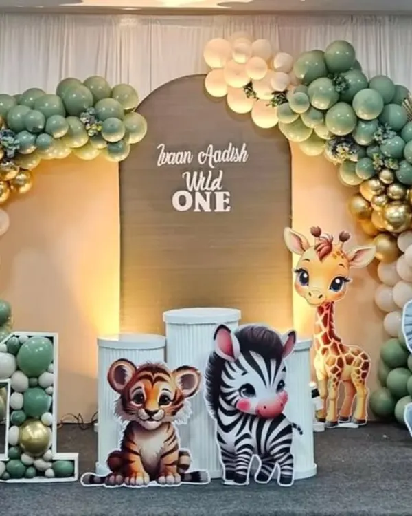 A chic 'Wild One' jungle safari theme for a first birthday. The setup includes a modern arched backdrop, a stylish balloon garland in sage green, white, and gold, and adorable cutouts of a lion, zebra, tiger, and giraffe.