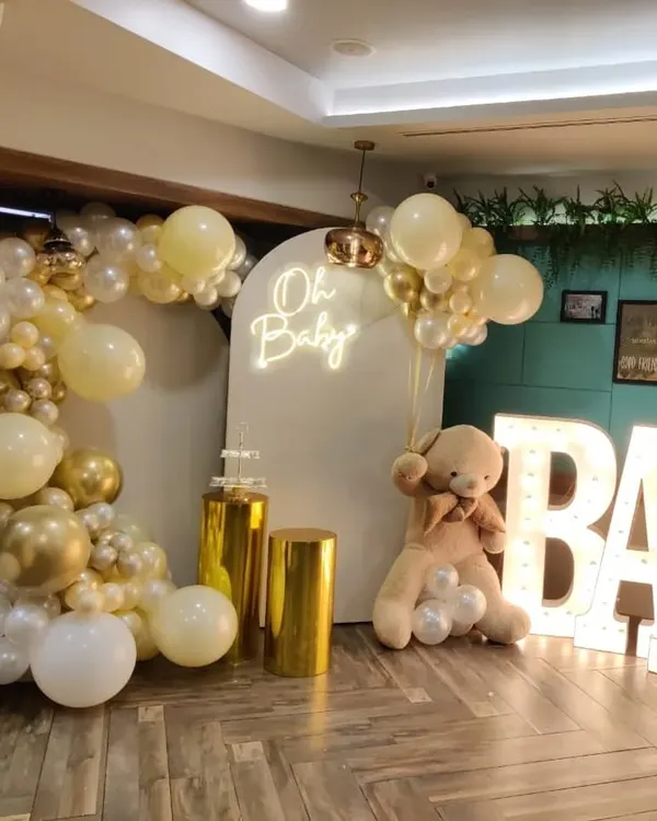 A full view of a classic "Oh Baby" shower setup. This popular theme includes a white and gold balloon garland, an arched backdrop with a neon sign, a large teddy bear prop, and light-up "BABY" letters.