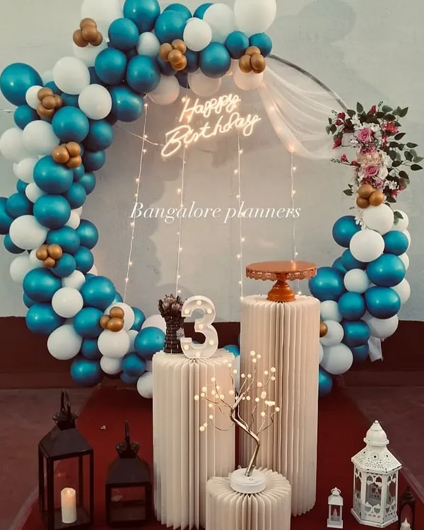 This full-circle balloon arch in shades of blue, white, and gold creates a sophisticated and festive photo corner. Paired with modern pedestals for the cake and a "Happy Birthday" neon sign, it's perfect for a stylish celebration.