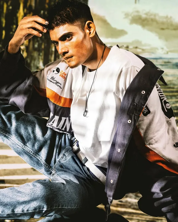 A relaxed yet stylish portrait. The projected beachside background and warm lighting create a casual, lifestyle feel that complements the modern, layered outfit.