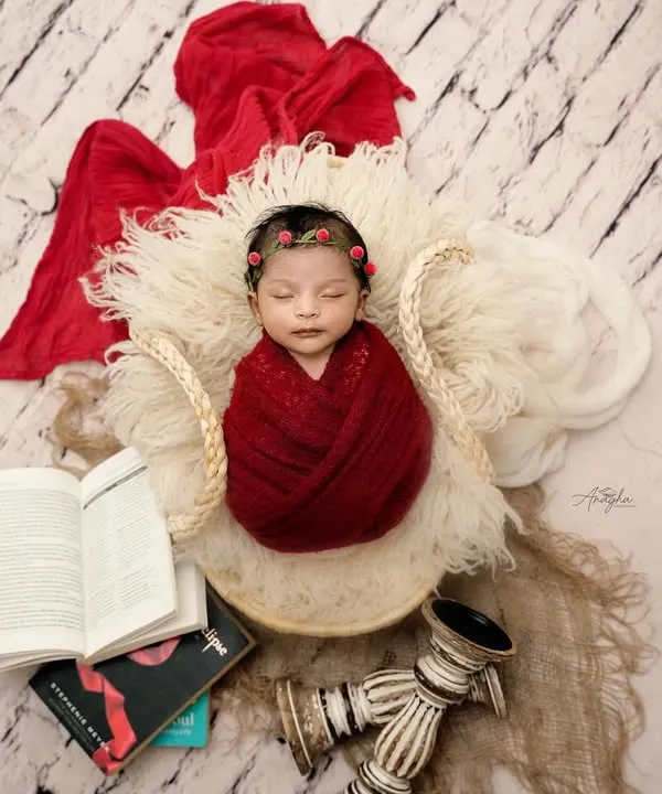 A beautifully composed overhead shot of a newborn wrapped in a rich red knit, sleeping in a basket surrounded by books and vintage decor.