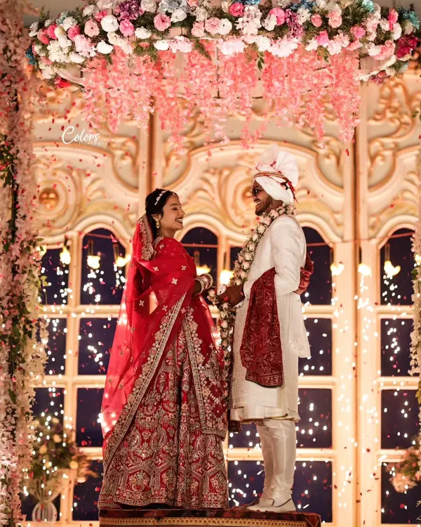 A breathtaking Varmala moment on an elevated stage, set against a stunning floral backdrop and a shower of petals. This is a true fairytale shot.