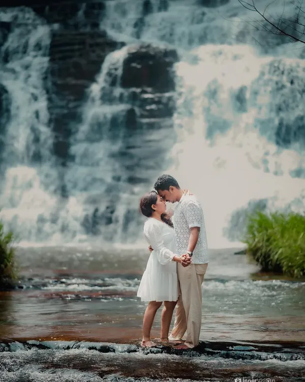A truly epic and romantic pre-wedding moment. The couple shares a kiss while standing on a rock in the middle of a river, with a magnificent waterfall cascading behind them.