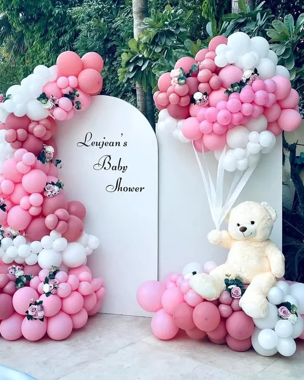 An elegant and sophisticated baby shower theme. This beautiful outdoor setup by the pool features a teddy bear holding a bouquet of pink and white balloons, with a matching organic balloon arch against a clean white backdrop.