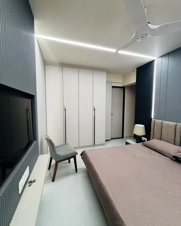 A wide view of a modern bedroom featuring a floor-to-ceiling white wardrobe and a contrasting dark grey fluted panel behind the TV. The linear ceiling lights provide clean, even illumination.