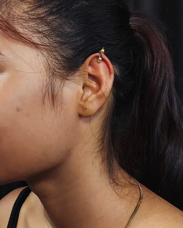 A close-up of a helix piercing on the upper ear cartilage with a decorative gold-tone barbell.
