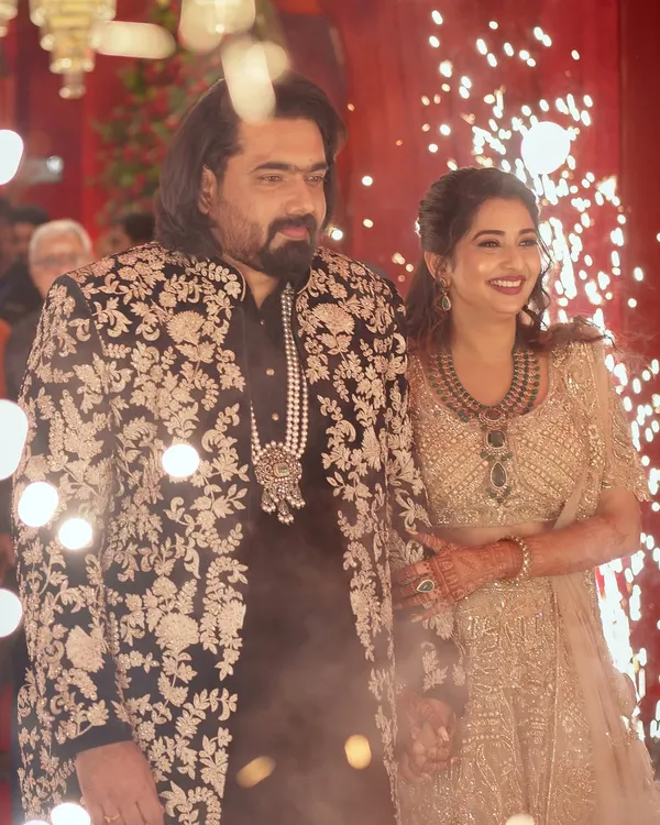 A grand entrance for a lifetime of smiles. The bride glows in a golden lehenga, perfectly complemented by the groom's black sherwani featuring intricate white floral embroidery.