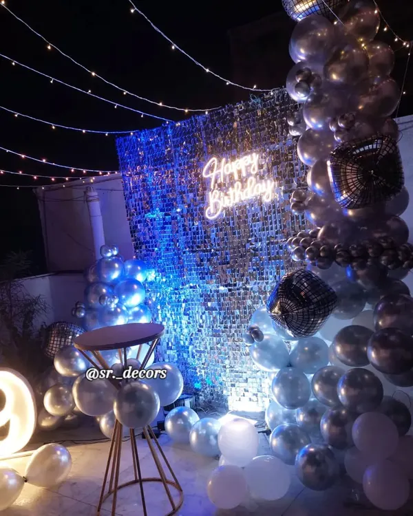 A glamorous rooftop party for a 29th birthday, featuring a silver shimmer wall, a balloon garland, fairy lights, and a glowing neon sign.