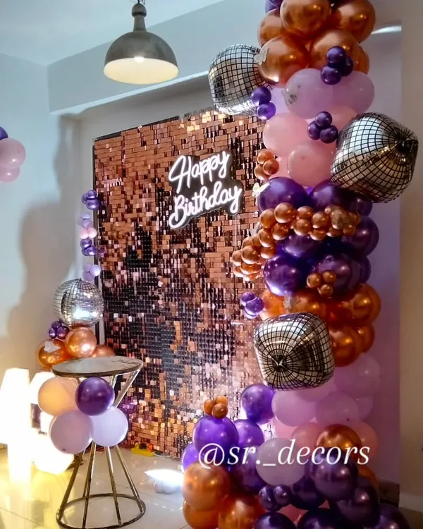 A stunning rose gold shimmer wall creates the perfect photo backdrop, complemented by a custom 'Happy Birthday' neon sign, a cascade of metallic and purple balloons, and sparkling disco balls for a truly glamorous party vibe.