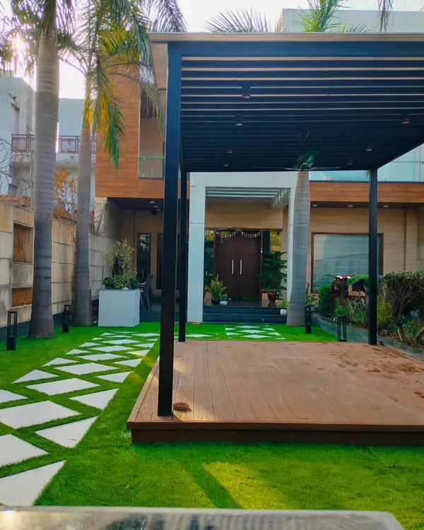 A completed front yard project in Dwarka, New Delhi, featuring a large wooden deck under a black pergola, surrounded by a manicured lawn and geometric pathway.