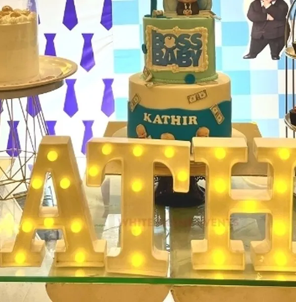 A close-up of custom light-up marquee letters spelling a name, placed on a glass table in front of a "Boss Baby" themed birthday cake.