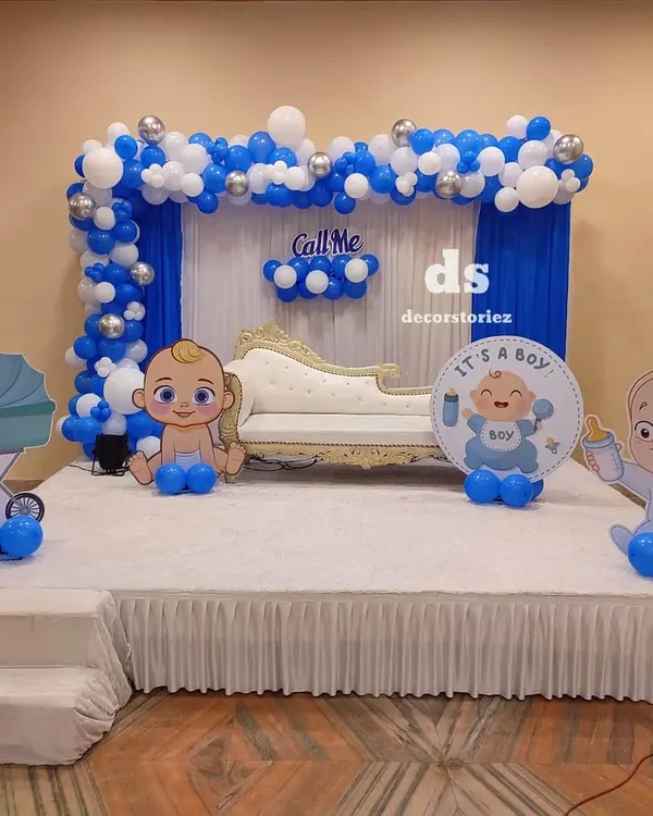 This stage decoration for a baptism ceremony features a blue and white balloon arch over drapes, with a comfortable sofa for the family. Themed cutouts and a circular sign add a personalized touch to the setup.