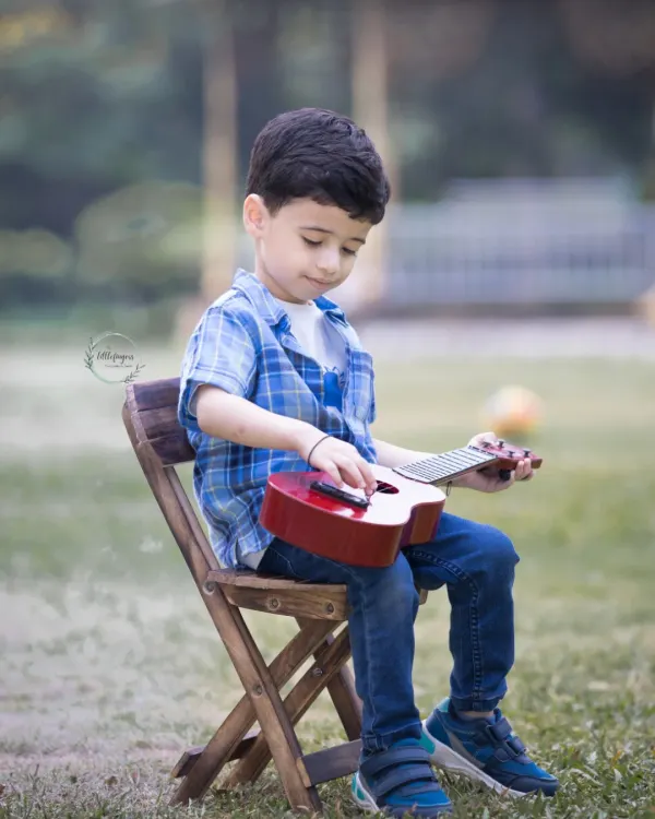 A little boy sits peacefully on a small wooden chair outdoors, focused on the toy guitar in his hands. A perfect portrait of childhood innocence and budding creativity.