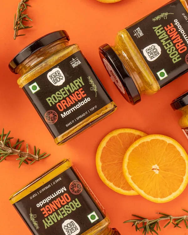 A dynamic flatlay for Pepper Wicks' Rosemary Orange Marmalade. The scattered jars, fresh orange slices, and rosemary sprigs clearly communicate the product's ingredients and flavor profile in a visually exciting way.