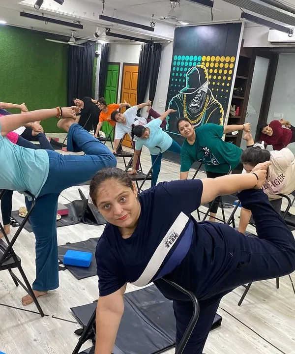 A full class of smiling faces enjoying a supported balancing pose in Chair Yoga. This practice is perfect for building stability and confidence at any age.