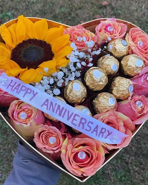 A heart-shaped anniversary box celebrating love with a bright sunflower, delicate pink roses, and a cluster of Ferrero Rocher chocolates. The personalized "Happy Anniversary" ribbon completes this thoughtful gift.