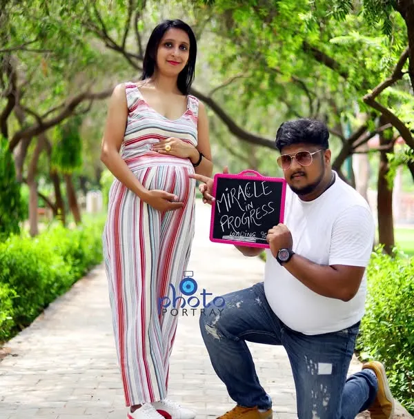 A creative announcement photo from a maternity shoot, with the father to be holding a sign that says "Miracle in Progress" while kneeling beside his partner.