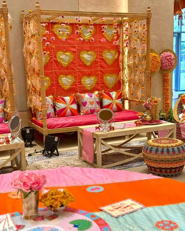 A festive seating arrangement for a Garba celebration, featuring a wooden four-poster canopy. The backdrop is a vibrant pink quilt with golden heart motifs, complemented by colorful custom cushions and traditional decor elements.