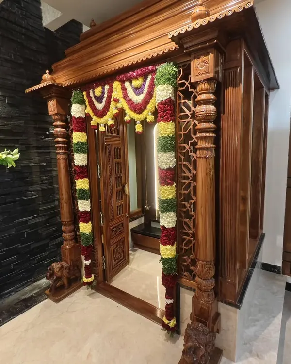 A stunning traditional wooden pooja room entrance, beautifully decorated with thick garlands of fresh flowers for a housewarming ceremony.