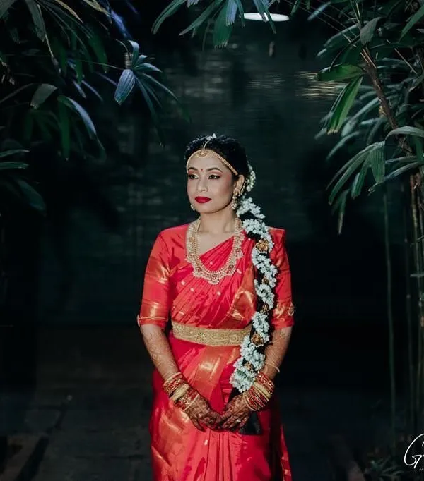 This is a classic South Indian bridal portrait. The makeup features a bold red lip and defined eyes, perfectly complementing her traditional red saree, temple jewelry, and jasmine-adorned braid for a timeless look.