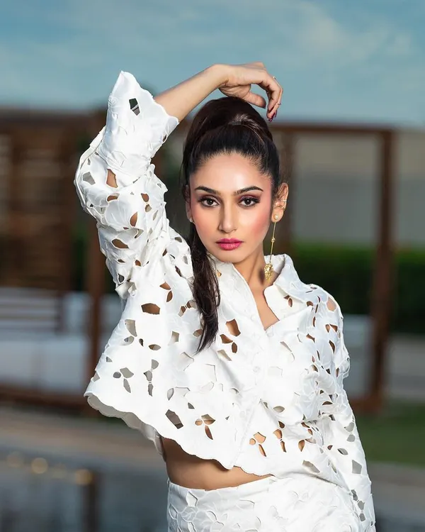A high-fashion outdoor portrait for a magazine. The pose and high ponytail hairstyle complement the structured, modern design of the white laser-cut outfit.