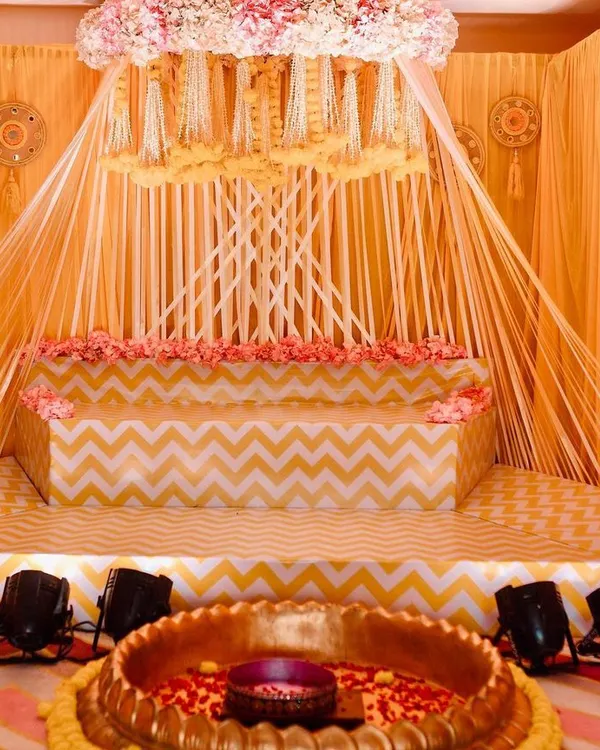 Here is the full view of the beautiful Haldi ceremony stage I designed. The setup features a canopy of hanging floral strings, a tiered seating area with a chevron pattern, and a large brass uruli in the front for the rituals.