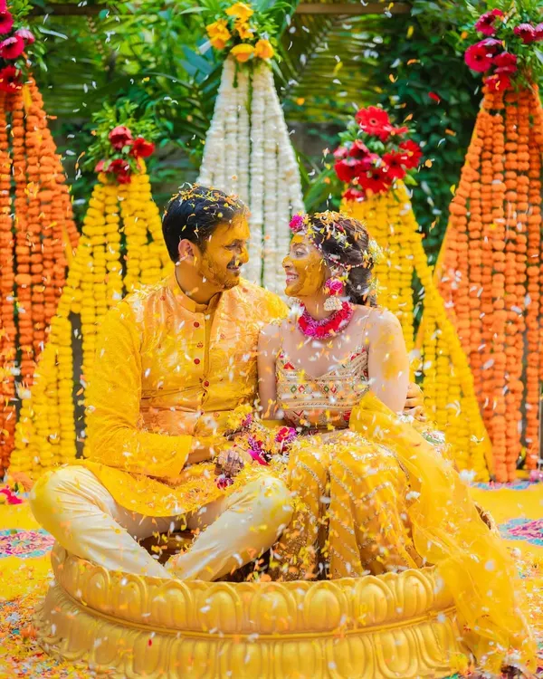 A picture-perfect Haldi moment, with the couple seated in a lotus-shaped tub, showered with flower petals against a vibrant marigold backdrop.