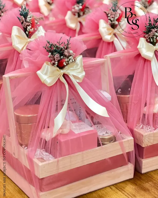 A beautifully arranged hamper in a rustic wooden crate, wrapped in delicate pink tulle and topped with a cream satin bow and dried flowers. This style is perfect for bridesmaid proposals, baby announcements, or any celebration deserving of a soft, elegant touch.