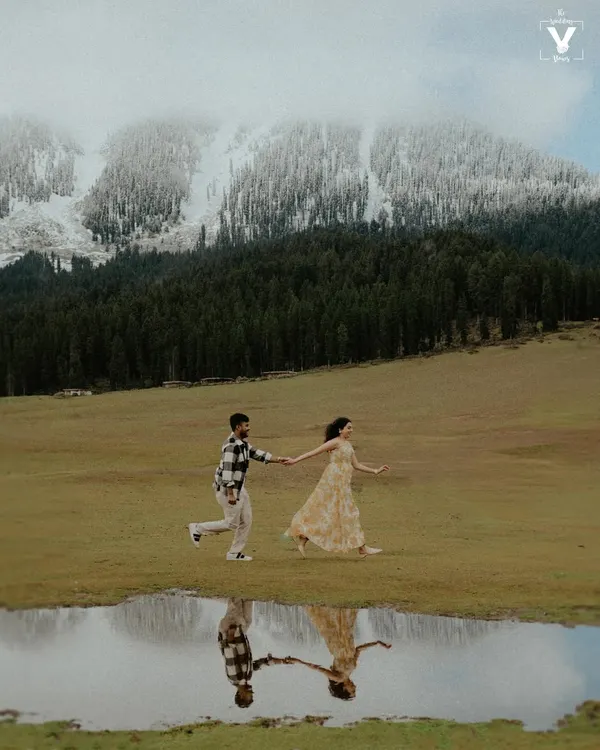 A couple runs hand-in-hand through a vast meadow in Kashmir, their reflection captured perfectly in a puddle, with snow-capped mountains in the background. This is the kind of cinematic pre-wedding photography we love to create.