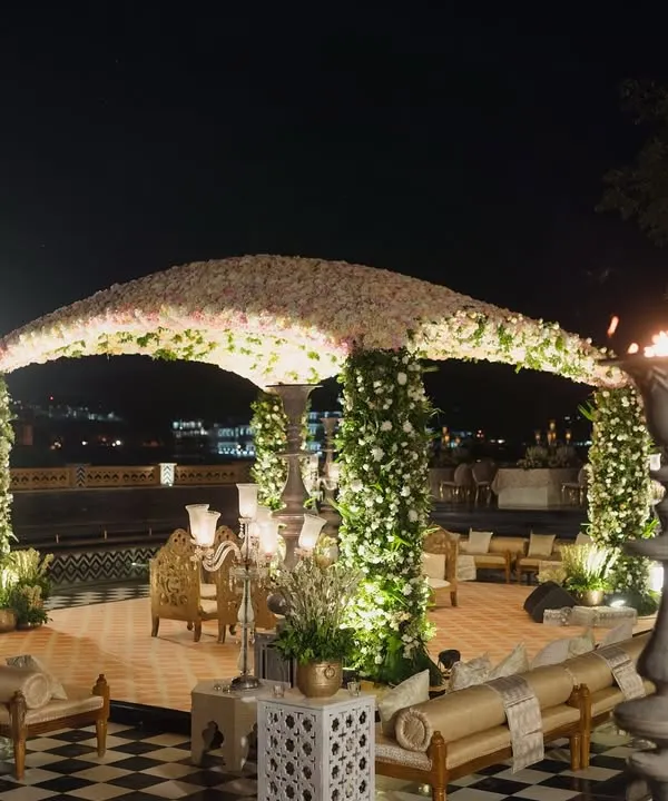 A stunning floral dome mandap set against the Udaipur city palace skyline at night. The structure is supported by pillars wrapped in white flowers, creating an open and airy feel for the wedding ceremony.