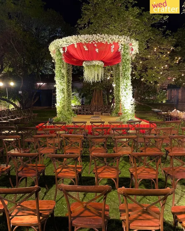 A bold and beautiful outdoor mandap at night, featuring pillars of greenery, a vibrant red canopy, and a chandelier of white flowers, surrounded by rustic wooden chairs.