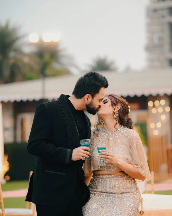 A candid moment from a cocktail party or reception. The couple shares a kiss while holding drinks, capturing the relaxed and celebratory vibe.