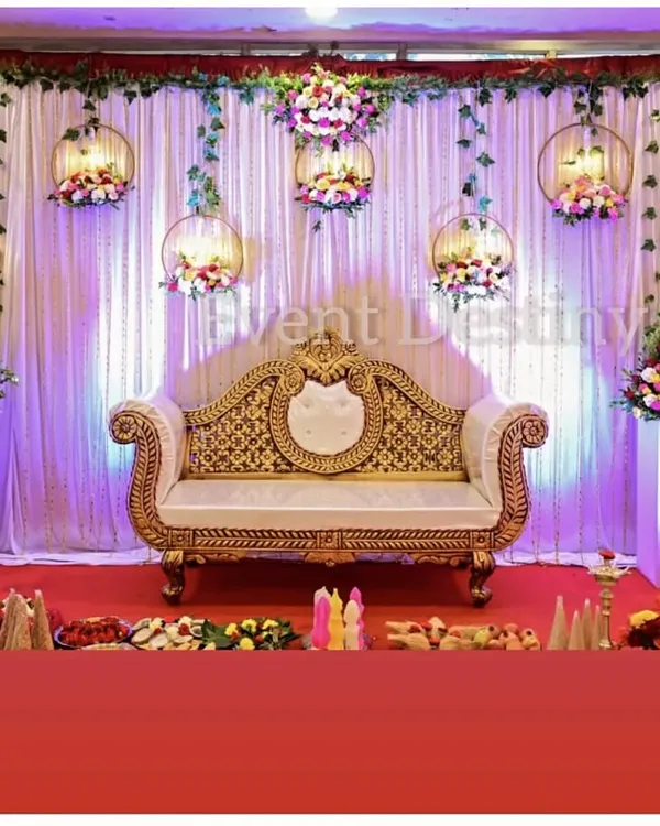 An elegant stage setup for an engagement or reception. The design features a white drape backdrop with hanging floral hoops and uplighting for a magical effect.