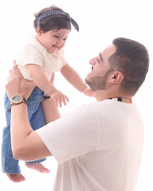 A classic father-daughter portrait against a clean white background. The father lifts his baby girl, capturing a moment of pure joy and connection in our bright and timeless style.