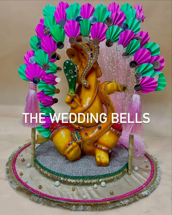 Welcome Bappa home with a custom mandap from us. This vibrant design features a Ganesha idol placed on a decorative platform, framed by a colorful arch made of pink and green paper fans with mirror work.