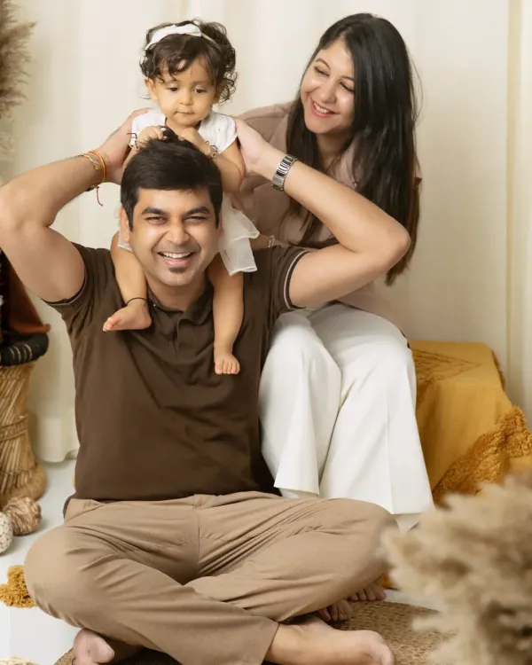 A playful studio family portrait where a father gives his daughter a shoulder ride, with mom looking on with a huge smile.