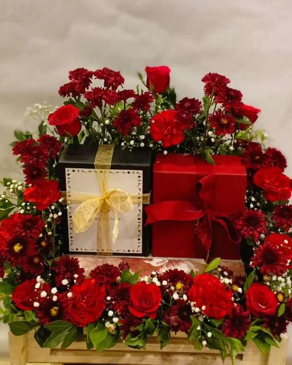 This luxurious hamper is a statement of pure elegance. It features two gift boxes nestled within a lavish arrangement of fresh red roses, carnations, and other blooms, perfect for a very special occasion.