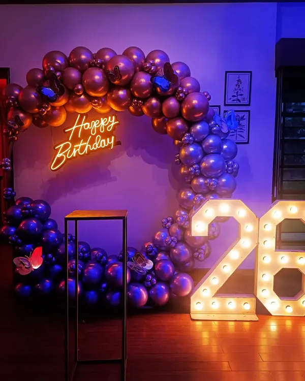 A complete birthday setup for a 26th celebration, featuring a full balloon ring in chrome purple and rose gold. The decor is enhanced with a custom 'Happy Birthday' neon sign and large, illuminated marquee numbers.