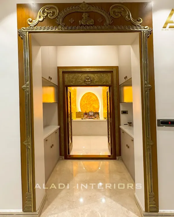 A grand entrance to a puja room, framed by an ornate, traditionally carved wooden archway. The inner doors feature intricate brass work, leading to a sanctum with a backlit onyx-style panel.